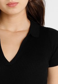 Top corto a maniche corte nero a coste con scollatura a V e colletto, realizzato in tessuto testurizzato. Il design presenta linee verticali sottili.