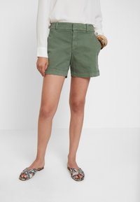 Zelené bavlněné šortky s přiléhavým střihem, které mají boční kapsy a hladký povrch, doplněné o vzorované otevřené sandály.