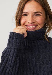 Navy blue turtleneck sweater with ribbed texture. The wearer is smiling, holding the collar. Features soft, thick fabric and a relaxed fit.
