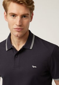 Polo shirt blu navy con colletto a righe bianche e piccolo logo ricamato a forma di bassotto sul lato sinistro del petto. Tessuto morbido e testurizzato, vestibilità classica.