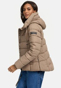 Béžová páperová bunda s kapucňou, s horizontálnym prešitím, logom na rukáve a zipsom vpredu. Nosená cez denim.
