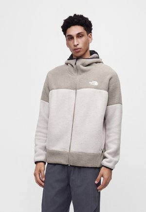 Jeune homme portant une veste polaire beige et gris clair zippée avec capuche et pantalon foncé, debout devant un fond uni.