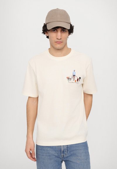 Kraft T-skjorte i kremfarge med korte ermer og grafisk trykk på venstre bryst. Kombinert med lyseblå jeans og en beiget cap.