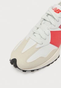 Białe i beżowe sneakersy z czerwonym logo "N", białymi sznurowadłami oraz czarną teksturowaną końcówką podeszwy na białym tle.