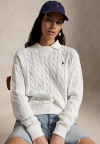 Ženska, ki nosi bel pulover z vzorcem pletenja, belo srajco z ovratnikom, mornarsko modro kapo in svetlo modre denim kratke hlače, sedi ob nevtralnem ozadju.