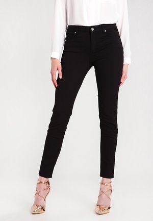 Mujer con pantalones negros ajustados, blusa blanca de manga larga y zapatos de tacón alto dorados rosados con tiras en el tobillo, de pie sobre un fondo blanco.