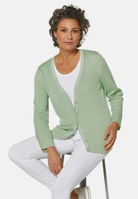 Hellgrüne gerippte Strickjacke mit V-Ausschnitt, langen Ärmeln und Knopfverschluss. Kombiniert mit weißen Hosen und vor einem schlichten Hintergrund.