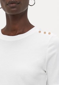 White sweatshirt with a crew neckline, soft texture, featuring three decorative gold-tone studs on the left shoulder.