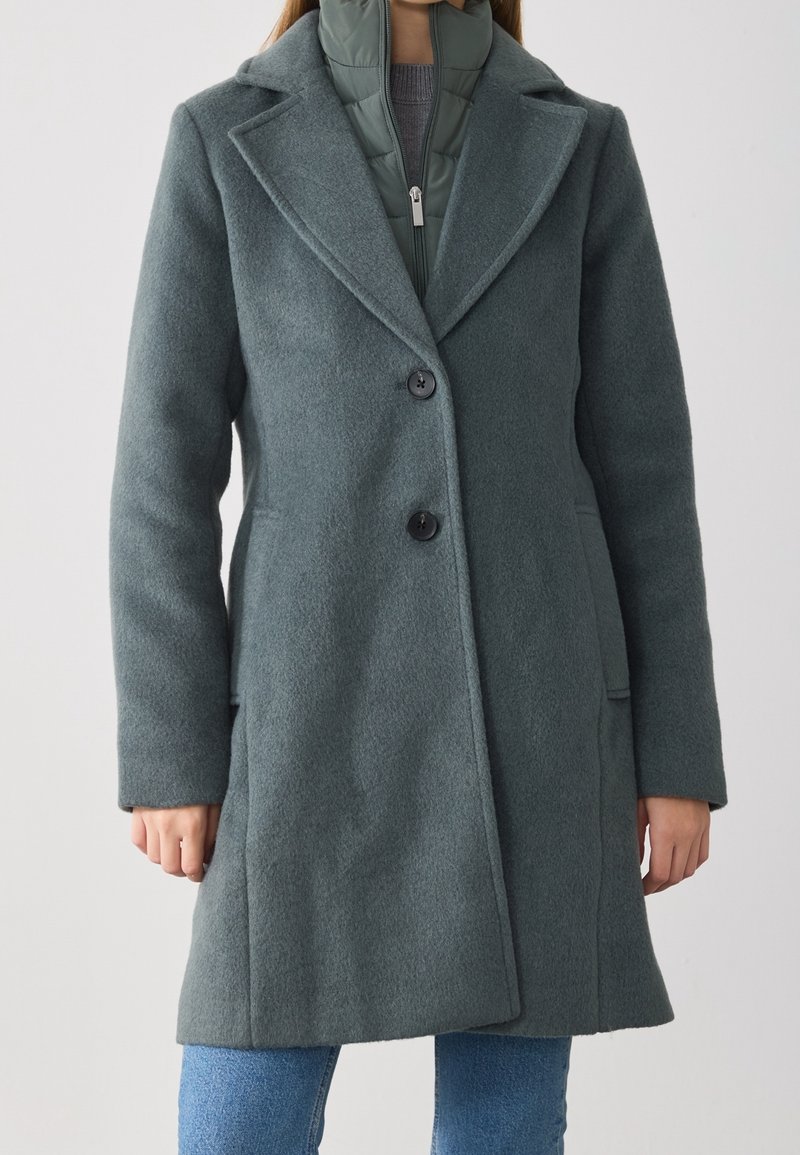 Femme portant un manteau en laine vert foncé boutonné par-dessus une veste matelassée verte et un jean bleu, debout devant un fond uni.