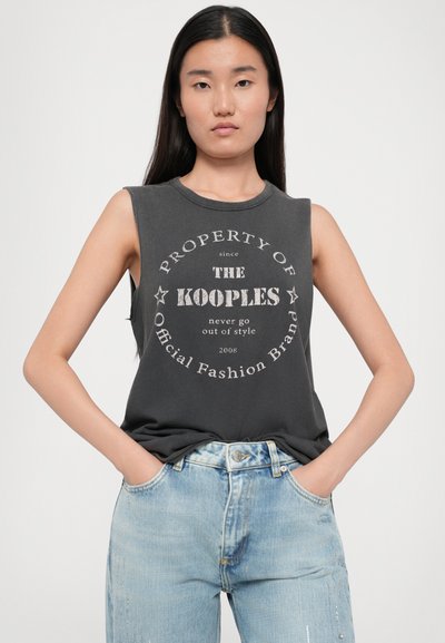 Mørk grå ermeløs t-skjorte med en rund grafikktrykk som står "PROPERTY OF THE KOOPLES," kombinert med lyseblå slitte jeans.