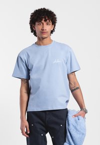 Camiseta de algodón azul claro con cuello redondo, mangas cortas y un pequeño logo blanco en el pecho. Textura suave, corte relajado, diseño minimalista.