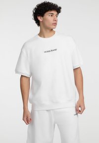 Valge üleriigne sweatshirt lühikeste varrukatega ja soonikitega, mille esiküljel on väike must "GUESS JEANS®" logo. Pehme tekstuur.