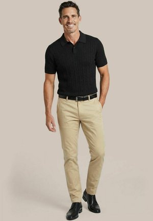 Lachende man die een zwart poloshirt met korte mouwen, beige broek, zwarte riem en zwarte nette schoenen draagt, staand met één hand in zijn zak.