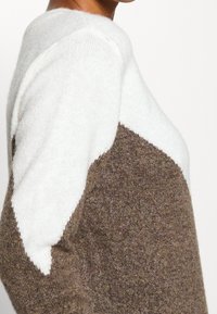 Stickad tröja med en vit övre del och en brun nedre del, prydd med ett chevronmönster. Mjuk textur, rund halslinning och långa ärmar.