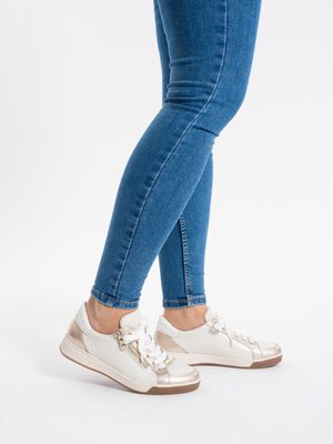Personne portant un jean skinny bleu et des baskets blanches avec des accents en or rose, debout jambes croisées sur un fond blanc.