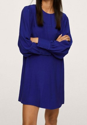 Kobieta ubrana w luźną, królewsko niebieską sukienkę z długim rękawem i mankietami na guziki, stojąca z założonymi rękami na tle jednolitego jasnego tła.