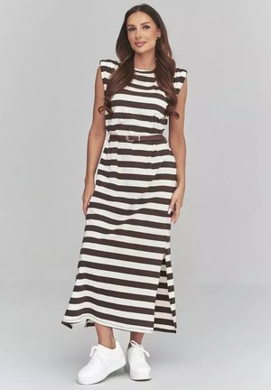 Kobieta stoi, ubrana w czarno-białą, poziomo paskowaną sukienkę bez rękawów, z brązowym paskiem i białymi sportowymi butami, uśmiecha się do aparatu.