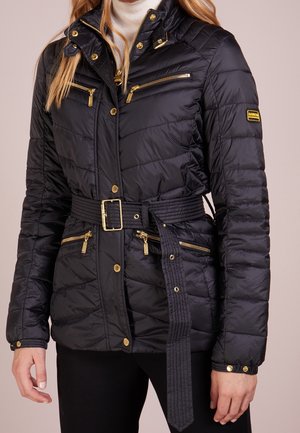 Zwarte gewatteerde jas met gouden ritsen, knopen en een riemgesp, gedragen door een persoon met lang blond haar, gecombineerd met een zwarte broek.