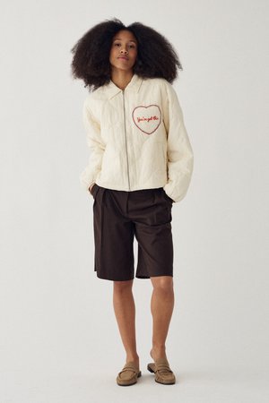 Frau mit natürlichem Haar, die eine cremefarbene, gesteppte Jacke mit einem Herz-Patch "You've got this", schwarze Shorts und beige Schlupfschuhe trägt und steht.