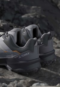 Ζεύγος γκρι και μαύρα παπούτσια τρεξίματος Adidas Terrex σε βραχώδες έδαφος, με επενδυμένους γιακάδες και υφασμένες εξωτερικές σόλες για καλύτερη πρόσφυση.