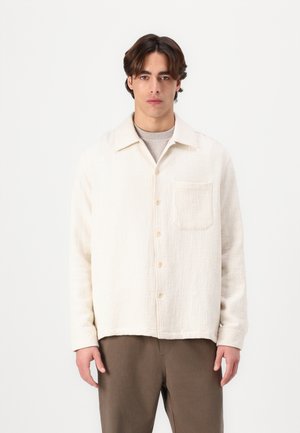 Licht beige overhemd met knopen en een gestructureerde stof, voorzien van een borstzak en een klassieke kraag, gedragen over een lichtgrijs shirt.