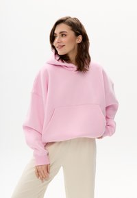 Pink übergroßer Hoodie mit einer Fronttasche, weiten Ärmeln und einer großen Kapuze mit Kordelzug. Gestylt mit beigen Hosen, vor weißem Hintergrund.