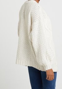 Krämfärgad stickad tröja med en strukturerad mönster, rundad halsringning och sänkta axlar, avslutad med ribbade ärmslut och nederkant.