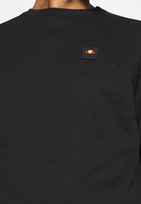 Sudadera negra de algodón con cuello redondo y un pequeño parche cuadrado con un logo en detalles rojos, amarillos y naranjas en el hombro.