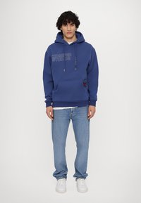 Felpa blu in cotone con tasca frontale, cappuccio con cordino e logo "KIDSUPER STUDIOS". Indossata sopra una maglietta bianca e abbinata a jeans azzurri chiari.