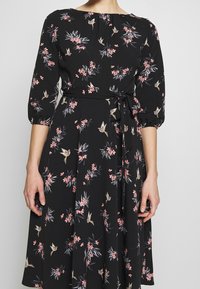 Svart blommig klänning med trekvartsärmar, med rosa blommor och fåglar, insvängd midja med ett knytband. Mjukt tyg med en luftig siluett.