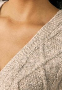 Beżowy sweter robiony na drutach z dekoltem w serek, mający teksturowany wzór i prążkowane krawędzie, podkreślający miękki, przytulny wygląd.