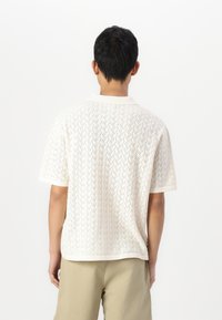 Polo branco de mangas curtas com padrão de malha em zigzag, colarinho e punhos de manga canelados. O tecido parece leve e respirável.