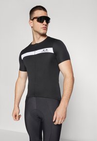 Schwarzes Radtrikot mit kurzen Ärmeln, weißem horizontalem Streifen und Logo. Enganliegendes Design kombiniert mit strukturierten schwarzen Radhosen.