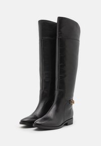Botas de cuero negras hasta la rodilla con una textura suave, punta cuadrada, tacón bajo en bloque y un acento de cadena en tono dorado en el costado.