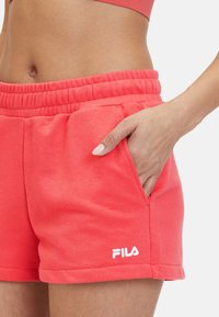 Pantaloni corti color corallo con vita elastica, tasche laterali e un logo bianco vicino all'orlo. Il tessuto sembra morbido e ha una texture liscia.
