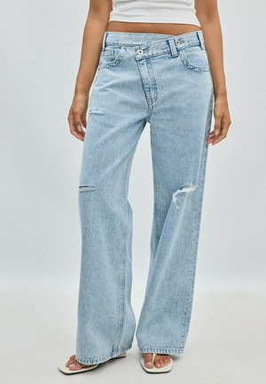 Femme portant un jean large bleu clair avec de petites déchirures sur un genou et une cuisse, associé à des sandales blanches à bout ouvert et un top court blanc.