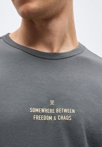 Κοντινό ενός ατόμου που φοράει γκρι πουκάμισο με το κείμενο «ΚΑΠΟΥ ΑΝΑΜΕΣΑ ΣΤΗΝ ΕΛΕΥΘΕΡΙΑ & ΤΟ ΧΑΟΣ» τυπωμένο με κρεμ αποχρώσεις στο στήθος.