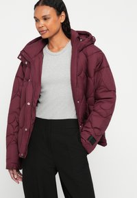 Giacca a vento bordeaux con motivo trapuntato, zip frontale, bottoni a pressione e cappuccio regolabile, indossata sopra un top grigio a coste e pantaloni neri.