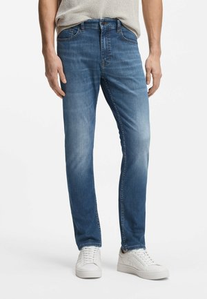 Mann trägt mittelblaue Denim-Jeans mit Verblassen an Oberschenkeln und Knien, kombiniert mit weißen Sneakers und einem beigen Stricktop.