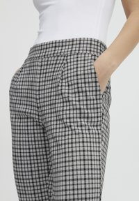 Kobieta w czarno-białych, kratkowanych spodniach z wysokim stanem, z bocznymi kieszeniami, ręką w kieszeni oraz w dopasowanej, białej bluzce bez rękawów.