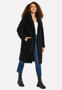 Cappotto nero, morbido, a pelo lungo con colletto a scarpetta, tasche frontali e bottoni. Indossato sopra una maglietta bianca e abbinato a jeans blu e stivali neri.