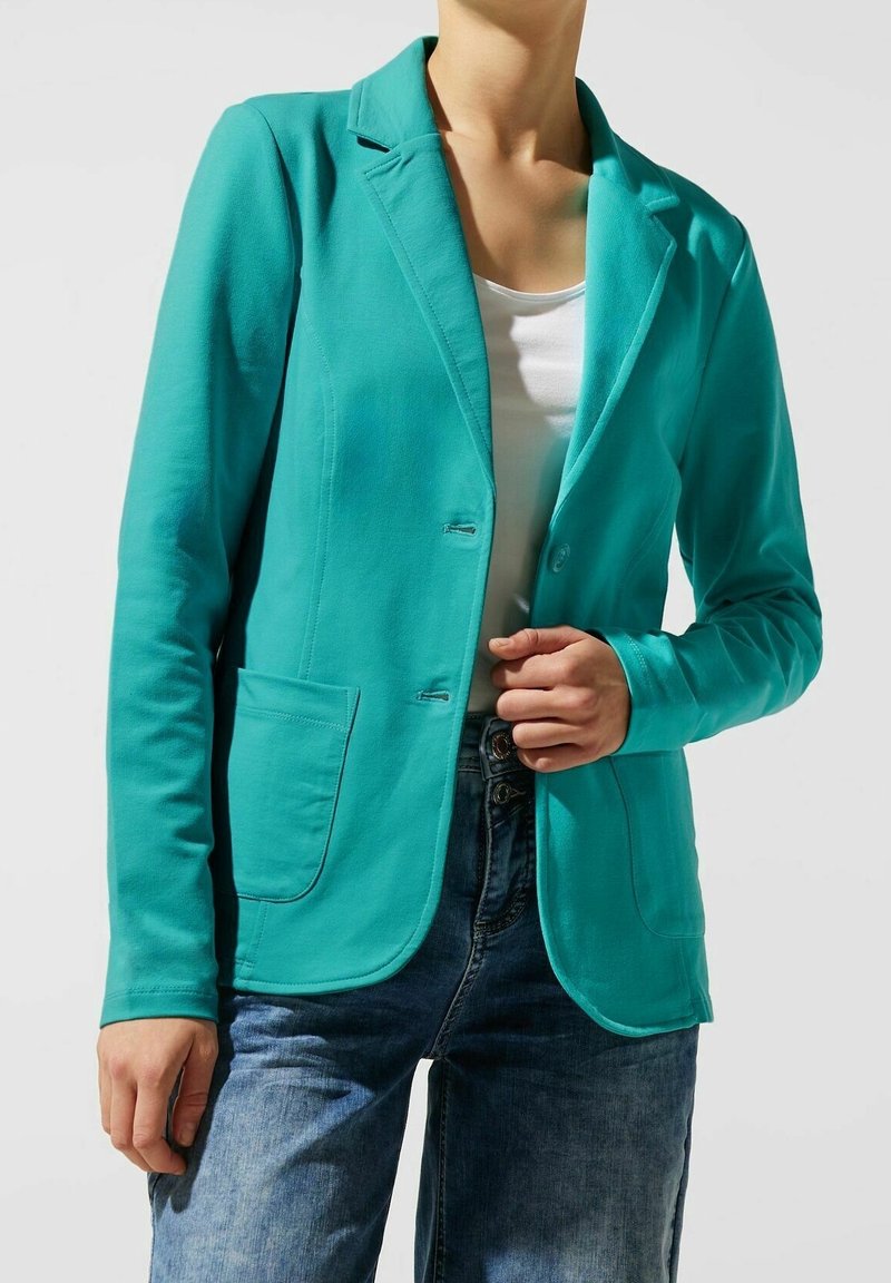 Blazer turquoise en tissu lisse et extensible. Il présente une fermeture à un bouton, deux poches avant et un col à revers.