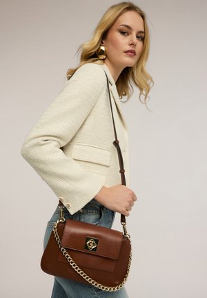 Femme blonde portant un blazer crème et un jean bleu, tenant un sac à main marron avec une chaîne et une fermeture dorées sur son épaule.