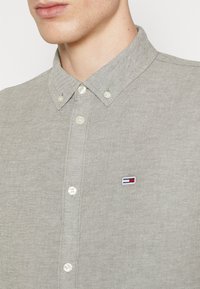 Ljusgrå knäppskjorta med spetsig krage, vita knappar och en liten broderad logotyp på vänster bröst. Strukturerad tyg.