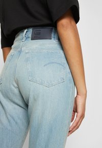 Jeans de ganga azul claro com cintura alta, apresentando dois bolsos traseiros e uma etiqueta de couro preto na cintura com um logo em relevo.