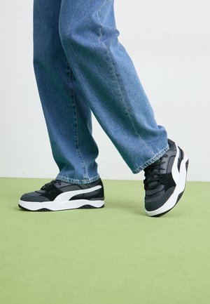 Personne portant un jean bleu et des baskets basses noires, blanches et grises, debout sur un sol vert avec un pied légèrement levé.
