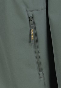 Grijze sportjack met een ritszak en een gestructureerde ritspull, en een label met de tekst "SUBLEVEL" in oranje. Glad stoffen afwerking.