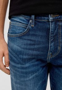 Dunkelblaue Jeans aus Denim mit klassischer Passform, sichtbaren Nähten, einer Fronttasche und einem Knopfverschluss. Glatte Textur.