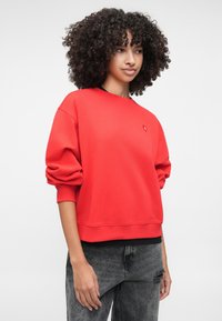 Κόκκινη αθλητική μπλούζα με στρογγυλή λαιμόκοψη, φαρδιές μανσέτες και ραμμένους μανίκια. Διαθέτει μια μικρή λεπτομέρεια λογότυπου στο στήθος και υφαντό ύφασμα.