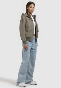 Veste à capuche grise avec fermeture éclair et deux poches avant. Portée par-dessus un haut blanc, associée à un jean large bleu clair et des baskets blanches.